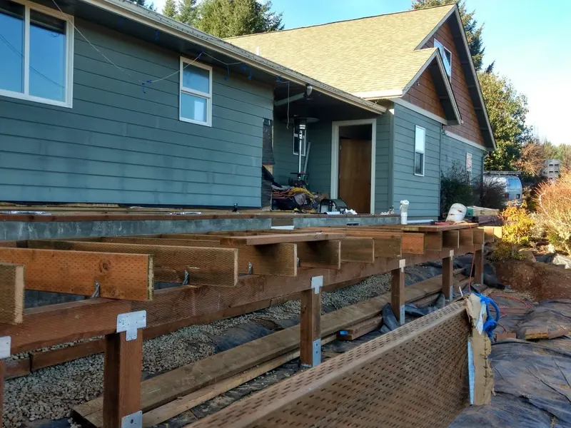 Ground-level view of heavy pressure-treated deck framing with galvanized hardware being built off a green-sided Oregon home by Red Umbrella.