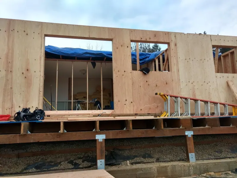 OSB-sheathed home addition wall with large picture-window and secondary window rough openings framed, showing the raised floor beam structure below.