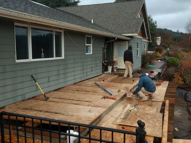 Workers complete subfloor framing on a raised home addition with an existing grey-green ranch home visible in the background.