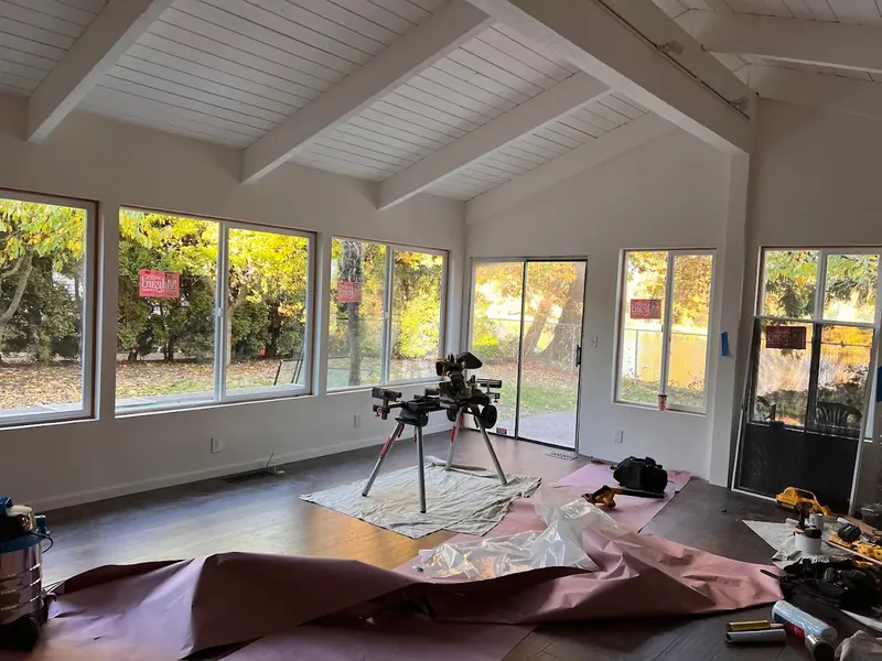 A nearly finished vaulted great room addition with white shiplap ceiling, exposed beams, and a panoramic window wall, under construction by Red Umbrella.