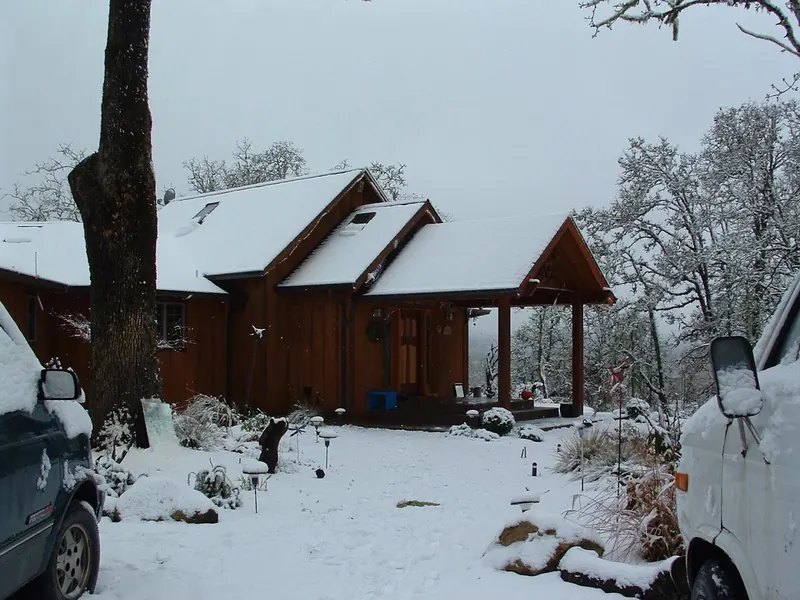 A stained cedar home with a covered gabled entry porch surrounded by snow-dusted oak trees in rural Oregon, built by Red Umbrella.