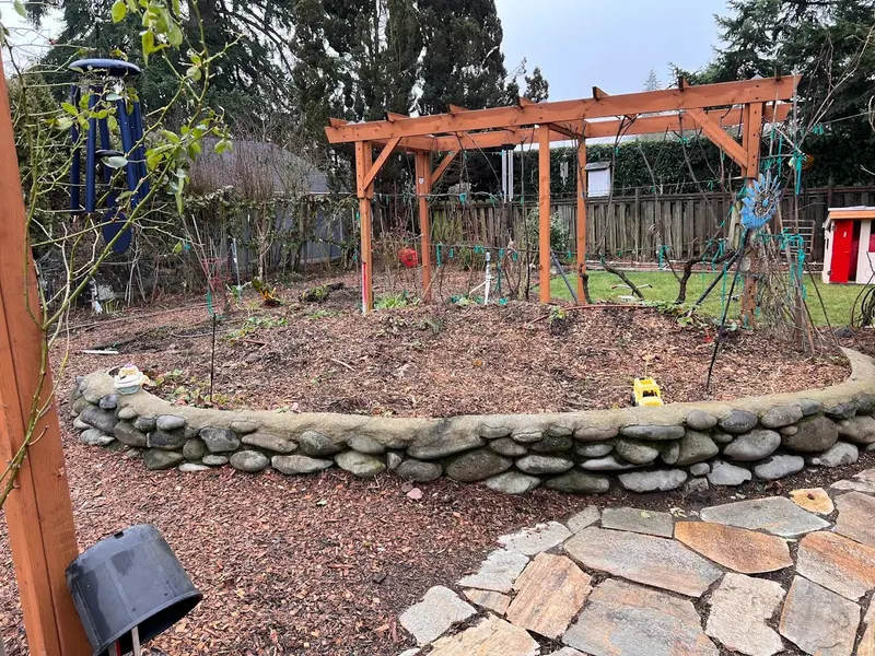 A curved dry-stacked fieldstone retaining wall surrounding a raised garden bed with a cedar pergola and flagstone patio, landscaping by Red Umbrella Eugene Oregon.