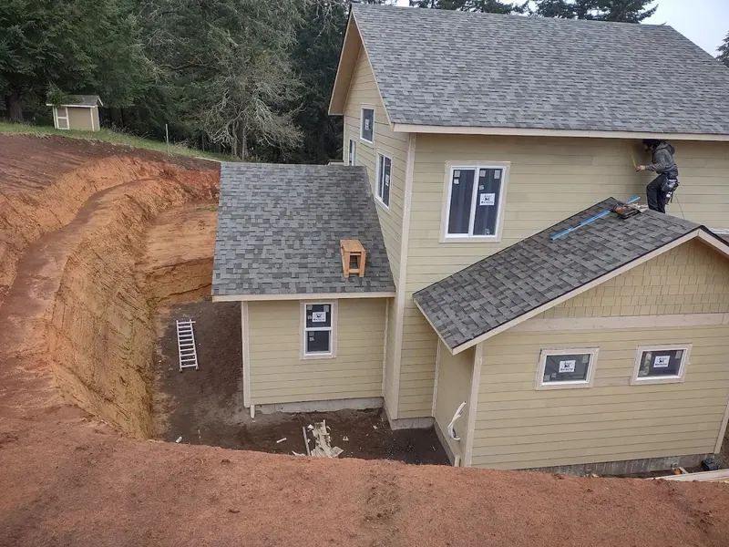 A two-story home addition with new sage green lap siding and charcoal asphalt shingles nearing completion on a wooded hillside lot, Red Umbrella Eugene Oregon.