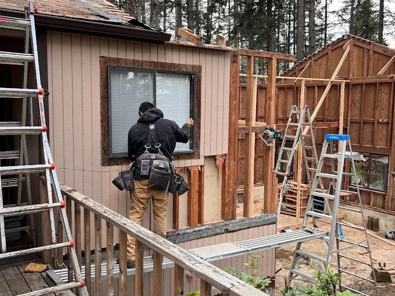 A worker installs a window in new addition framing adjacent to an existing house with board-and-batten siding, construction by Red Umbrella Eugene Oregon.