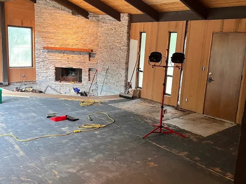 Exposed concrete subfloor in a mid-century great room during renovation by Red Umbrella, showing cedar ceiling, dark timber beams, and stacked-stone fireplace.